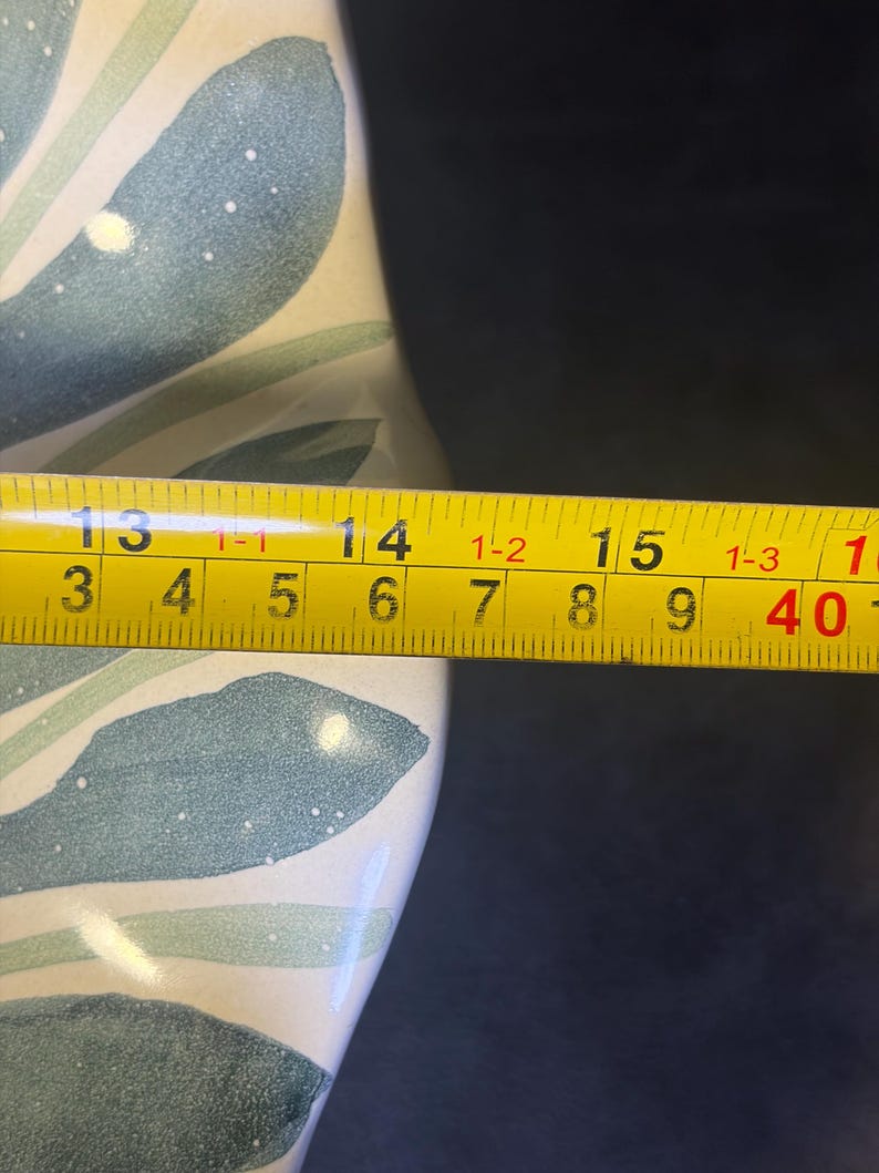 Op de afbeelding: Een gedeelte van een keramische vaas met een geschilderd bladmotief in tinten blauw en groen op een cr&egrave;mekleurige achtergrond. Een gele meetlint toont een lengte van ongeveer 35,5 cm.