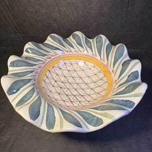 Op de afbeelding: Een decoratieve keramische kom met een geschulpte rand. De kom heeft een witte basis met een patroon van blauwgroene bladeren en roze stippen rond de rand. Het midden heeft een gele ring en een geometrisch ontwerp.