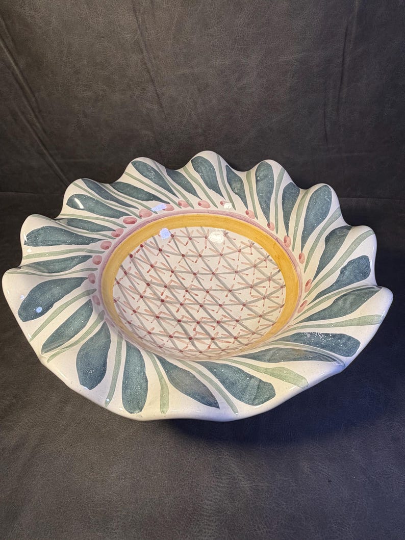 Op de afbeelding: Een decoratieve keramische kom met een geschulpte rand. De kom heeft een witte basis met een patroon van blauwgroene bladeren en roze stippen rond de rand. Het midden heeft een gele ring en een geometrisch ontwerp.