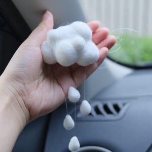 May include: A white felt cloud car charm with dangling teardrops. The cloud is held in a hand, with the car's interior visible in the background. The charm is designed to hang from a car's rearview mirror.