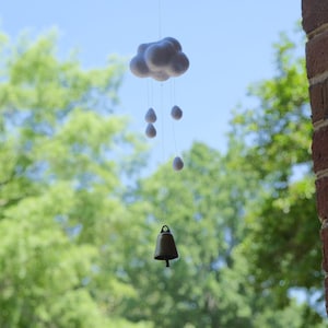 May include: A whimsical mobile featuring a fluffy, gray cloud with hanging teardrop-shaped rain drops. A small, bronze-colored bell hangs below. The mobile is suspended against a backdrop of a bright blue sky and green foliage.