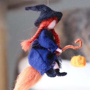 Può includere: Bambola di strega in feltro fatta a mano con trecce di filo rosso, indossa un vestito blu e un cappello nero, in sella a una scopa arancione. Una piccola zucca arancione è appesa alla scopa. Decorazione di Halloween.