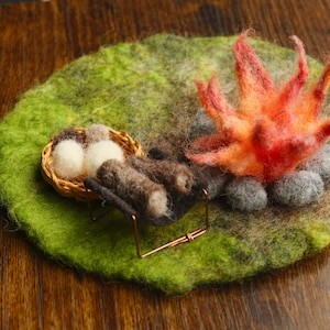Può includere: Una scena di falò in feltro con una base erbosa verde e gialla. La scena comprende un falò in miniatura con fiamme arancioni e rosse, pietre grigie e un piccolo cestino con uova bianche. I tronchi marroni poggiano su un piccolo supporto di metallo.
