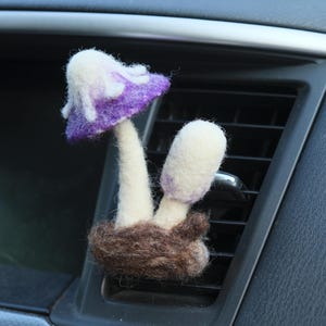 May include: A purple and white felt mushroom car air freshener hanging from the air vent of a car.