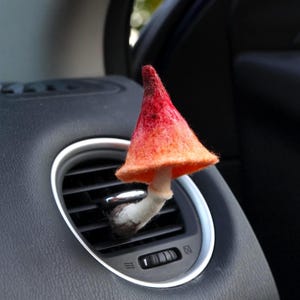 May include: A felt mushroom air freshener with a red and orange cap and a white stem, inserted into a car's air vent. The mushroom is a whimsical, decorative item.