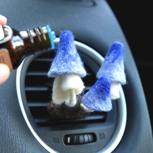 May include: Two felt mushroom car air fresheners with blue caps and white stems are placed in a car vent. A hand is holding a brown bottle, possibly essential oil, near the mushrooms. The car interior is dark gray.