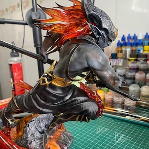 Puede incluir: Figura detallada de un guerrero demoníaco en una pose dinámica, empuñando una espada. La figura presenta un tono de piel gris, armadura negra con detalles dorados y cabello y detalles de color naranja fuego. La base es una estructura rocosa similar a la lava.