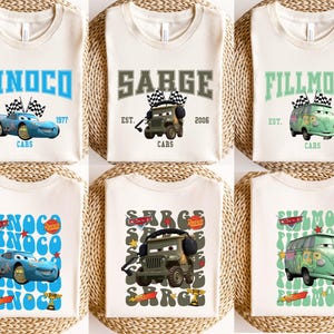 May include: Cream-colored t-shirts featuring cartoon car graphics and text. Designs include "DINOCO," "SARGE," and "FILLMORE," with additional text like "EST. 1977" and "CARS." The shirts are displayed on a woven surface.