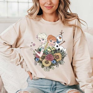 Pu&ograve; includere: Una felpa color crema con una stampa grafica di Elsa e Anna di Frozen, insieme a Olaf, circondato da fiori.