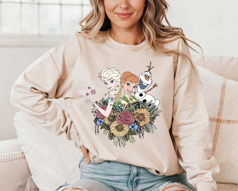Pu&ograve; includere: Una felpa color crema con una stampa grafica di Elsa e Anna di Frozen, insieme a Olaf, circondato da fiori.
