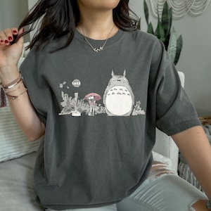 May include: Dark gray t-shirt featuring a graphic of a large, gray cartoon character with a white belly, standing next to a person holding a pink umbrella. The design includes a moon and foliage.