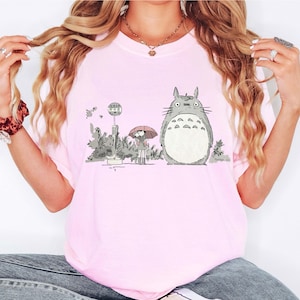 May include: Light pink t-shirt featuring a graphic of Totoro from My Neighbor Totoro. The illustration shows Totoro standing next to a bus stop with a girl holding an umbrella. The shirt is a soft, comfortable material.