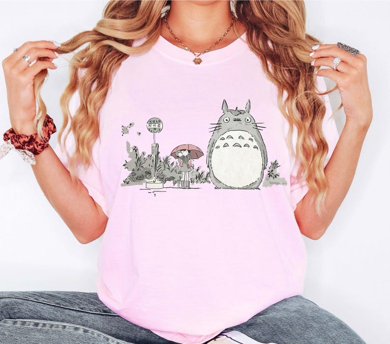 May include: Light pink t-shirt featuring a graphic of Totoro from My Neighbor Totoro. The illustration shows Totoro standing next to a bus stop with a girl holding an umbrella. The shirt is a soft, comfortable material.