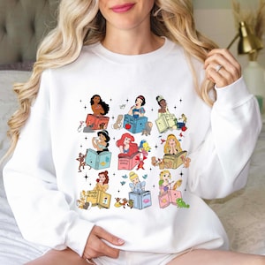 May include: White sweatshirt with a colorful graphic print of Disney princesses reading books. Each princess is holding a book with their name on the cover.