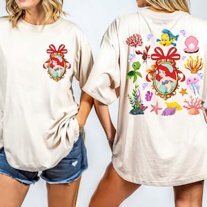 May include: Two white t-shirts with a cartoon image of Ariel from The Little Mermaid. The front shirt has a single image of Ariel in a gold frame with a red bow. The back shirt has multiple images of Ariel, sea creatures, and seashells.