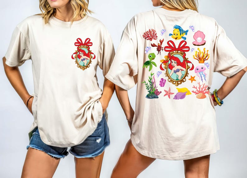 May include: Two white t-shirts with a cartoon image of Ariel from The Little Mermaid. The front shirt has a single image of Ariel in a gold frame with a red bow. The back shirt has multiple images of Ariel, sea creatures, and seashells.
