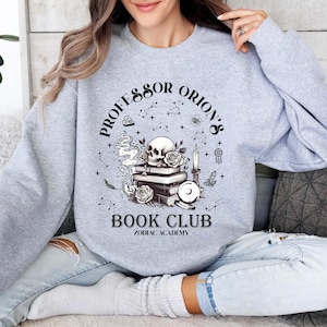 May include: A light grey sweatshirt with a graphic design featuring a skull, roses, books, and the text "Professor Orion's Book Club Zodiac Academy". The design includes constellations and other mystical elements.