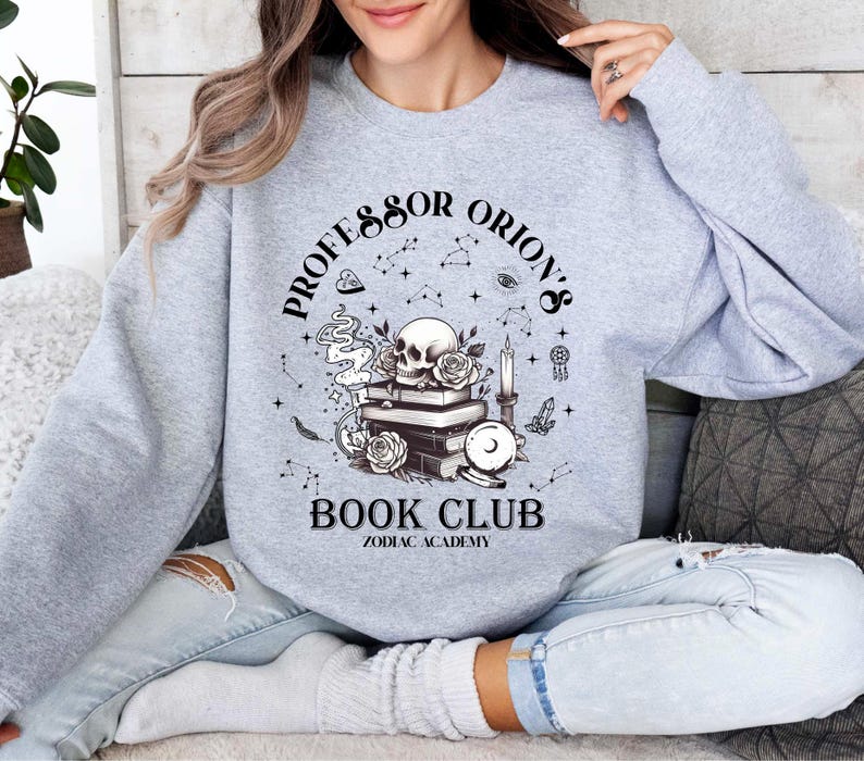 May include: A light grey sweatshirt with a graphic design featuring a skull, roses, books, and the text "Professor Orion's Book Club Zodiac Academy". The design includes constellations and other mystical elements.