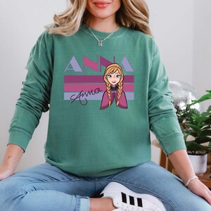 May include: A teal green sweatshirt with a graphic of Anna from Frozen. The design features the name "ANNA" in lavender and pink, with a cartoon image of Anna and her name in cursive below.