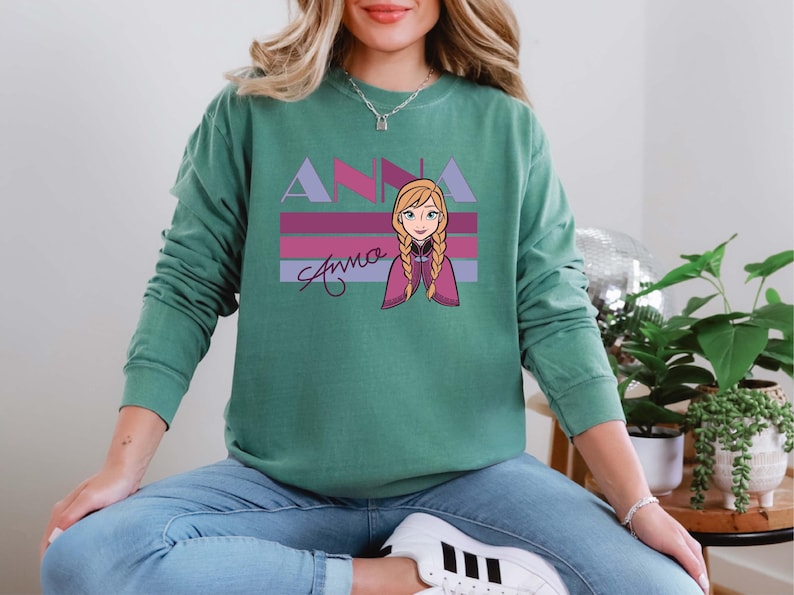 May include: A teal green sweatshirt with a graphic of Anna from Frozen. The design features the name "ANNA" in lavender and pink, with a cartoon image of Anna and her name in cursive below.