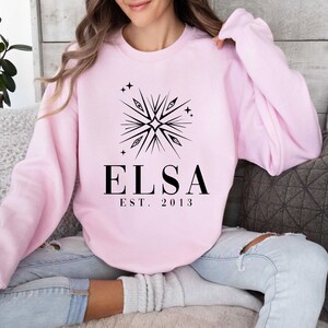 Puede incluir: Sudadera rosa con un dise&ntilde;o de copo de nieve negro y el texto "Elsa Est. 2013".