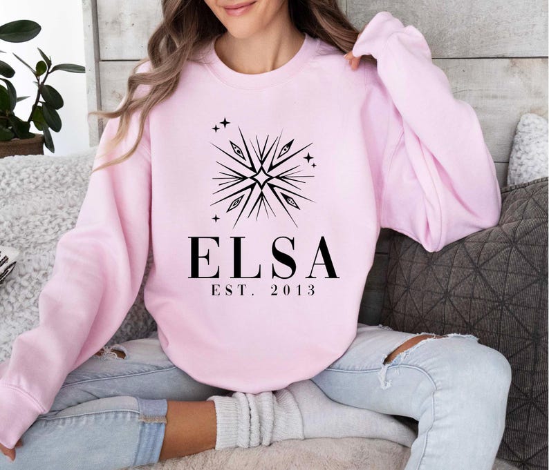 Puede incluir: Sudadera rosa con un dise&ntilde;o de copo de nieve negro y el texto "Elsa Est. 2013".
