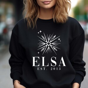 Puede incluir: Sudadera negra con un dise&ntilde;o de estrella blanca y el texto "ELSA EST. 2013" impreso en la parte delantera.