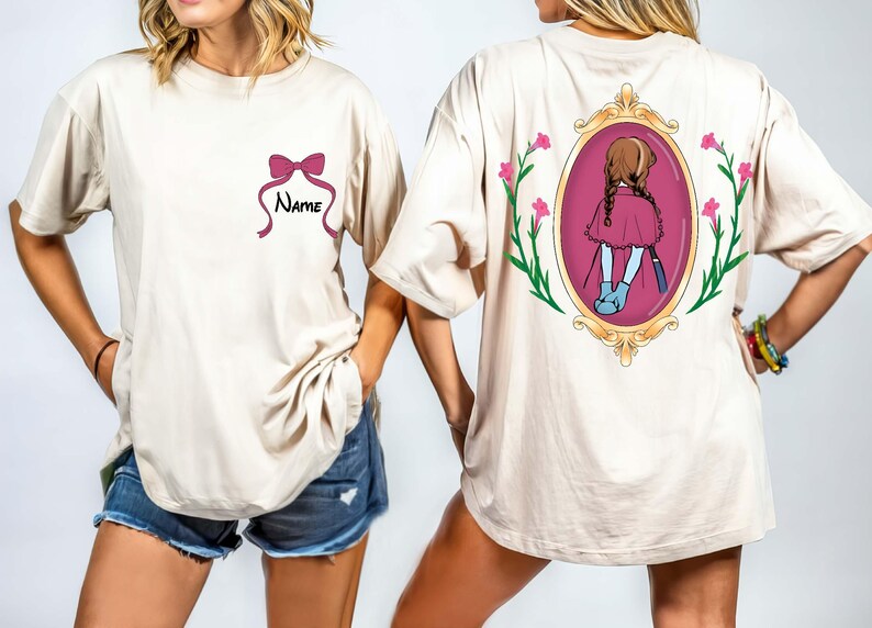 May include: Beige t-shirt with a pink bow and the word "Name" on the front. The back of the shirt features a framed illustration of a girl in a pink dress. Pink flowers and green stems surround the frame.