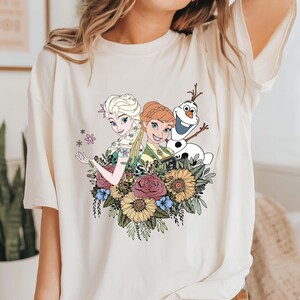 May include: A white t-shirt with a graphic print of Elsa and Anna from Frozen, along with Olaf. The characters are surrounded by flowers in shades of pink, yellow, and green.