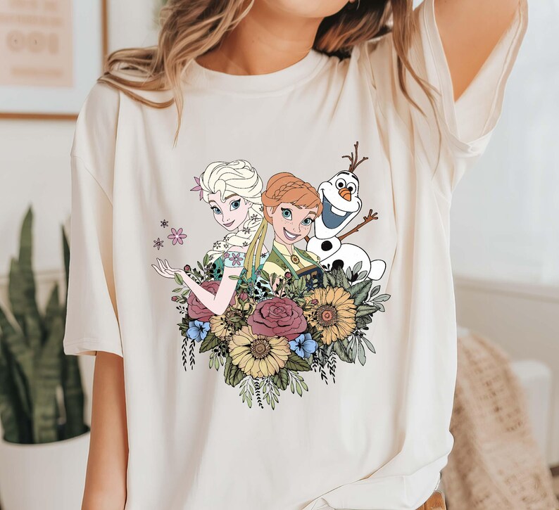 May include: A white t-shirt with a graphic print of Elsa and Anna from Frozen, along with Olaf. The characters are surrounded by flowers in shades of pink, yellow, and green.