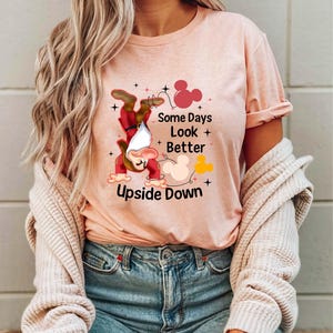 May include: A light pink t-shirt featuring a cartoon gnome design. The gnome is upside down with the text "Some Days Look Better Upside Down". The design includes red, white, and yellow accents.