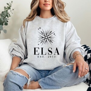 Puede incluir: Una sudadera gris con un dise&ntilde;o gr&aacute;fico negro de un copo de nieve con una estrella en el centro y el texto "ELSA EST. 2013".
