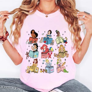May include: Pink t-shirt featuring a colorful graphic of various Disney princesses reading books. Each princess is depicted with her respective book title, surrounded by small animals and stars. The shirt is a light pink color.