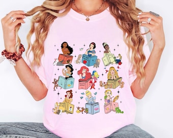 May include: Pink t-shirt featuring a colorful graphic of various Disney princesses reading books. Each princess is depicted with her respective book title, surrounded by small animals and stars. The shirt is a light pink color.