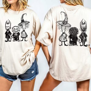 Pu&ograve; includere: T-shirt beige con un'illustrazione a fumetti in bianco e nero di tre personaggi sul davanti e sul retro. Il design include una strega, un personaggio con la testa rotonda e un personaggio con le corna. La maglietta ha maniche corte e una vestibilit&agrave; ampia.