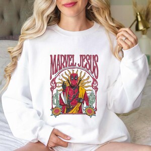 May include: White sweatshirt with a vintage-style graphic of Deadpool in a red and black suit with the text "Marvel Jesus" above him.