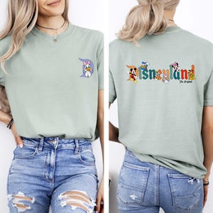 May include: Sage green t-shirt featuring a Daisy Duck graphic on the front and the Disneyland logo with Disney characters on the back. The text "The Original" is below the logo. The shirt is a short-sleeved, crew neck style.