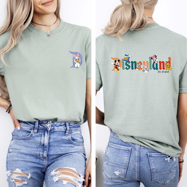 May include: Sage green t-shirt featuring a Daisy Duck graphic on the front and the Disneyland logo with Disney characters on the back. The text "The Original" is below the logo. The shirt is a short-sleeved, crew neck style.