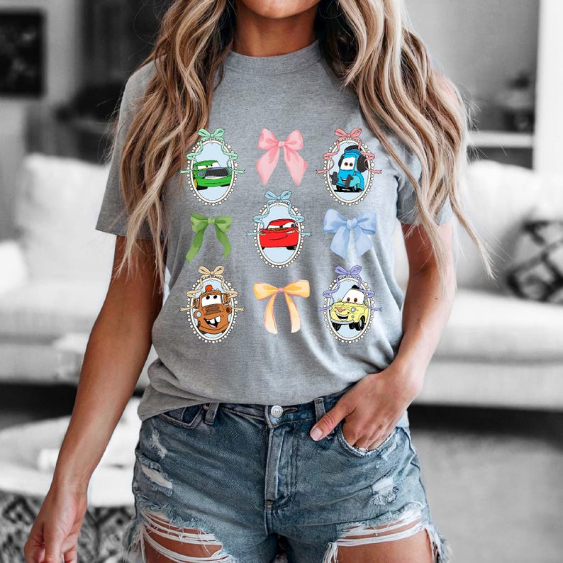 May include: A gray t-shirt featuring cartoon car characters in oval frames with colorful bows. The shirt has a crew neck and short sleeves. The cars are in various colors, including green, red, and blue.