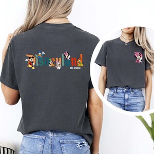 May include: Dark gray t-shirt featuring the word "Disneyland" in colorful letters with cartoon characters. The back of the shirt has Mickey Mouse, Donald Duck, Daisy Duck, Minnie Mouse, and Goofy. The text "The Original" is below the word Disneyland. A smaller image of Minnie Mouse is on the front.