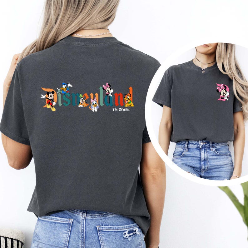 May include: Dark gray t-shirt featuring the word "Disneyland" in colorful letters with cartoon characters. The back of the shirt has Mickey Mouse, Donald Duck, Daisy Duck, Minnie Mouse, and Goofy. The text "The Original" is below the word Disneyland. A smaller image of Minnie Mouse is on the front.