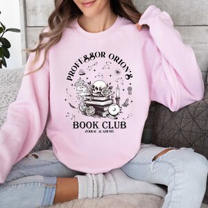 May include: Pale pink sweatshirt featuring the text "Professor Orion's Book Club Zodiac Academy" in black. The graphic includes a skull, books, roses, and celestial elements. The sweatshirt has long sleeves.