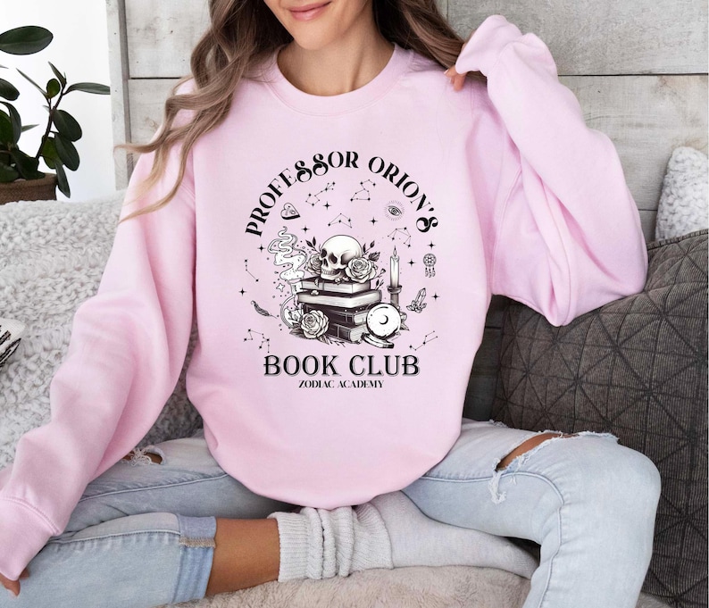 May include: Pale pink sweatshirt featuring the text "Professor Orion's Book Club Zodiac Academy" in black. The graphic includes a skull, books, roses, and celestial elements. The sweatshirt has long sleeves.