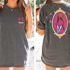 May include: Dark gray t-shirt with a pink bow and the name "Name" printed on the front. The back features a framed illustration of a girl in a red dress and blue boots. The frame is gold with pink flowers.