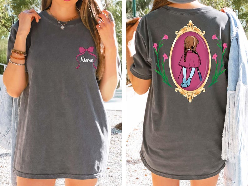 May include: Dark gray t-shirt with a pink bow and the name "Name" printed on the front. The back features a framed illustration of a girl in a red dress and blue boots. The frame is gold with pink flowers.