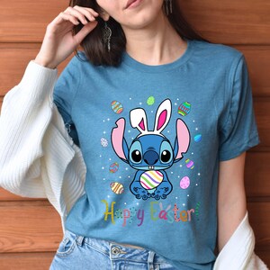 May include: A blue t-shirt with a cartoon Stitch wearing bunny ears and holding an Easter egg. The shirt has a colorful Easter egg pattern and the text "Happy Easter!"