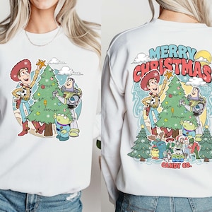 Puede incluir: Sudadera blanca con un gr&aacute;fico colorido de Woody, Buzz Lightyear y un extraterrestre decorando un &aacute;rbol de Navidad. La parte trasera de la sudadera tiene el texto "MERRY CHRISTMAS" en rojo y azul, con personajes y &aacute;rboles adicionales.