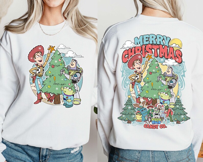 Puede incluir: Sudadera blanca con un gr&aacute;fico colorido de Woody, Buzz Lightyear y un extraterrestre decorando un &aacute;rbol de Navidad. La parte trasera de la sudadera tiene el texto "MERRY CHRISTMAS" en rojo y azul, con personajes y &aacute;rboles adicionales.