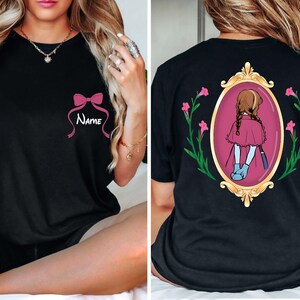May include: Black t-shirt featuring a pink bow with the word "Name" in white script. The back of the shirt displays a framed illustration of a girl in a pink cape, surrounded by flowers. A stylish and personalized apparel item.