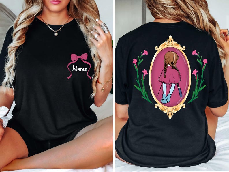 May include: Black t-shirt featuring a pink bow with the word "Name" in white script. The back of the shirt displays a framed illustration of a girl in a pink cape, surrounded by flowers. A stylish and personalized apparel item.